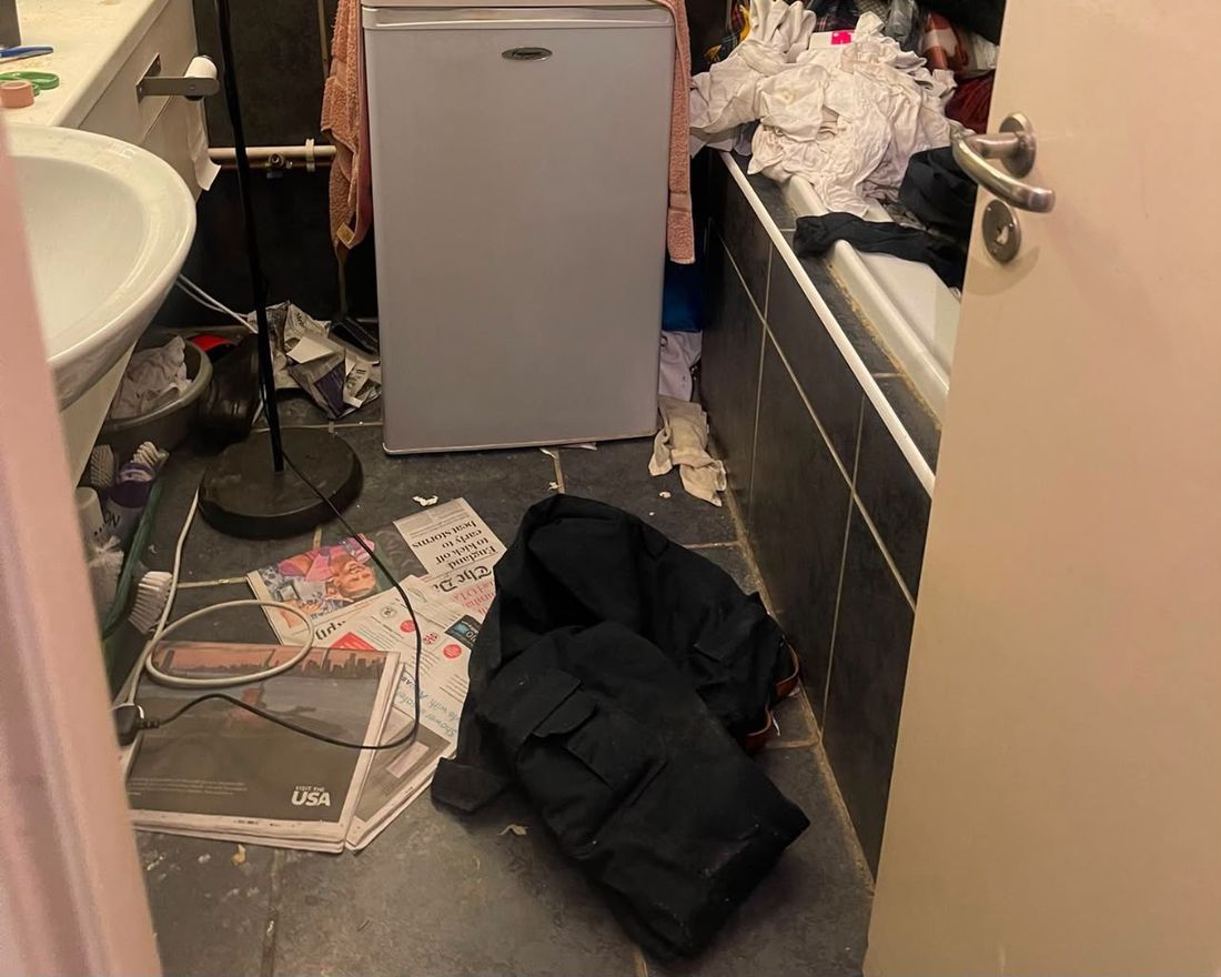 Bathroom before cleaning: a fridge, newspapers, clothes and rubbish covering the floor